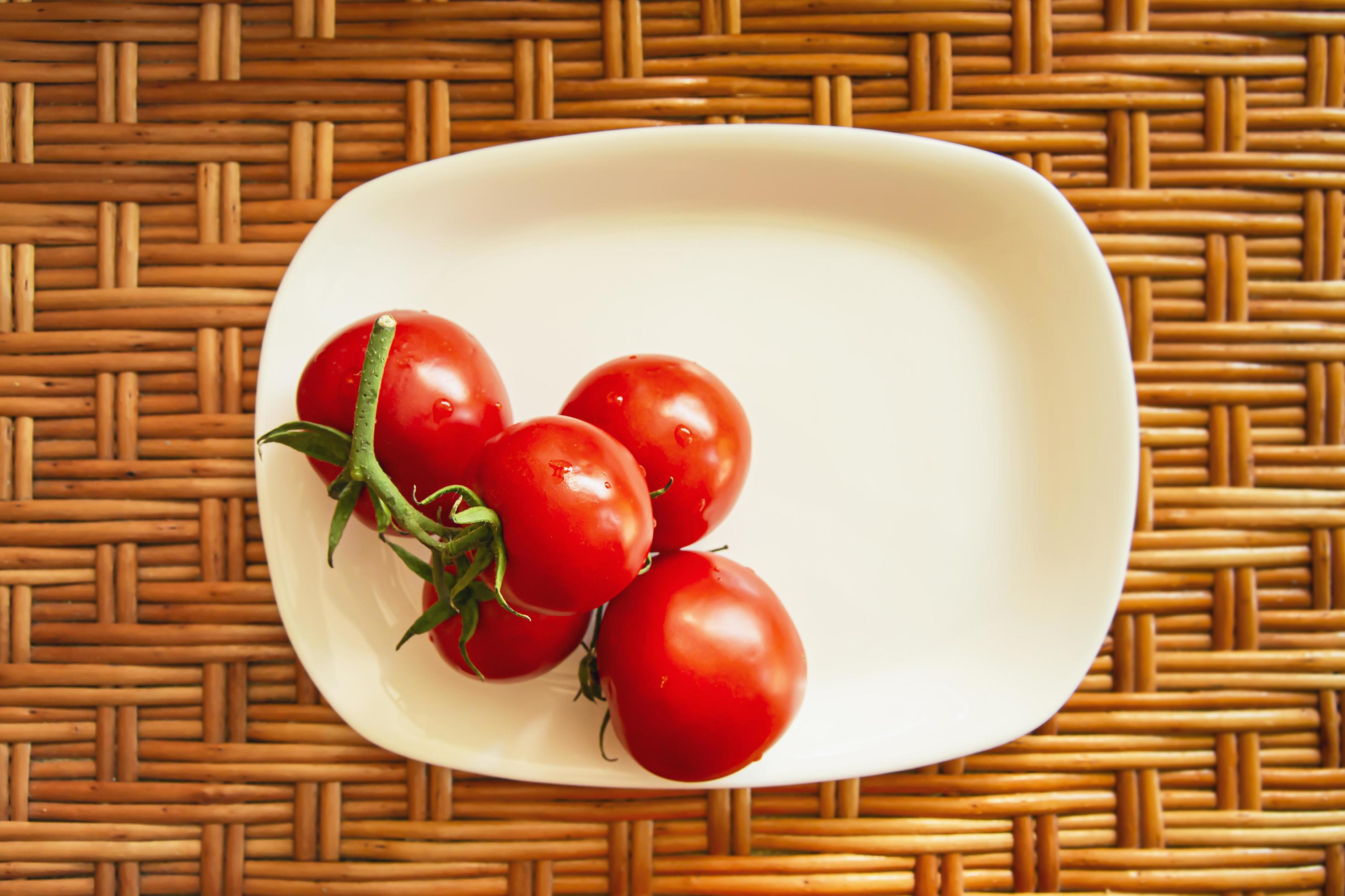 A cluster of red cherry tomatoes on a white flat rectangular plate stand on a wooden wicker surface. View from above. Life style composition. Stock Free