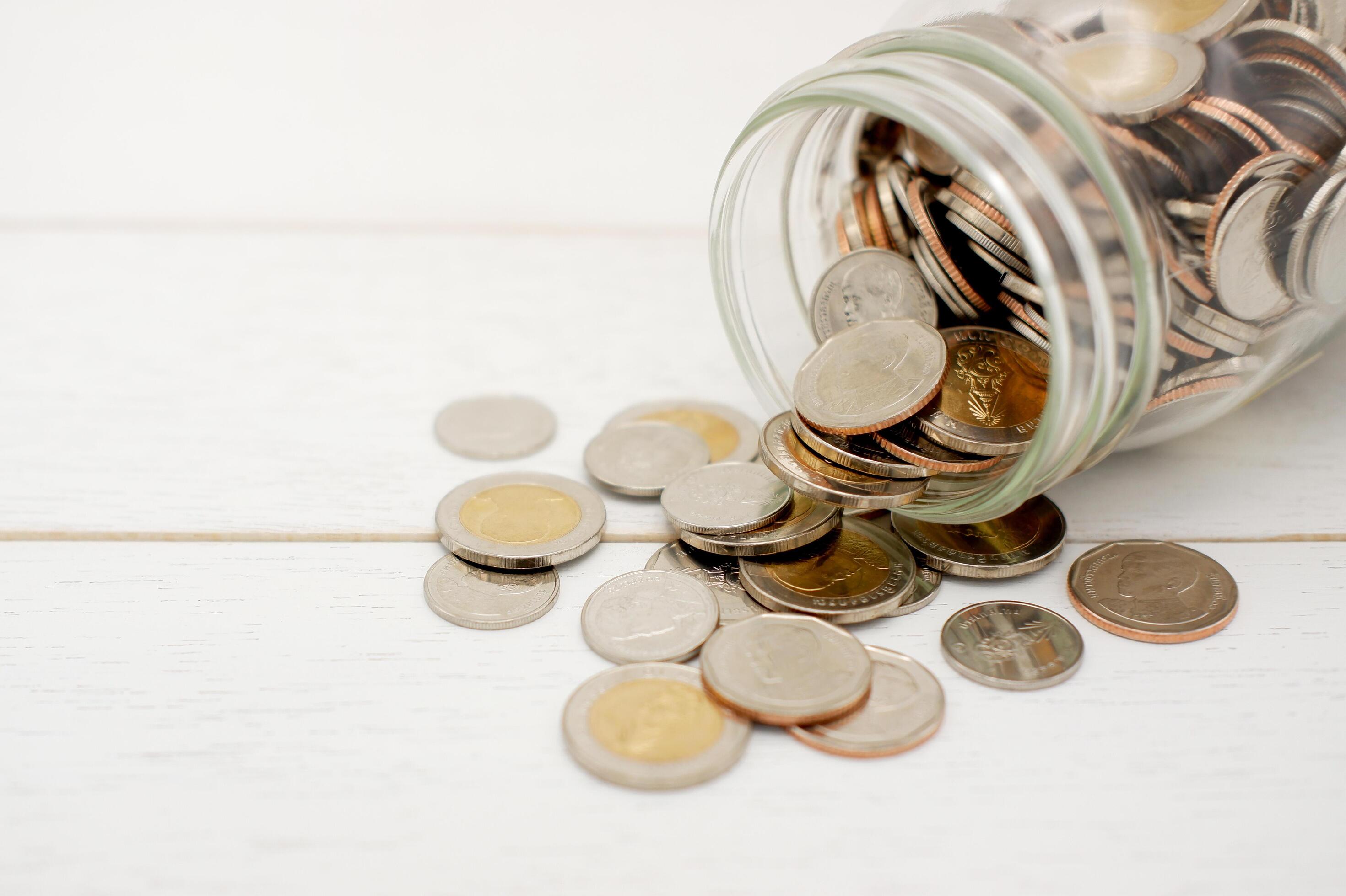 New Thai Baht coins spilling from a glass jar, on white wooden table. Business and finance concept. Stock Free