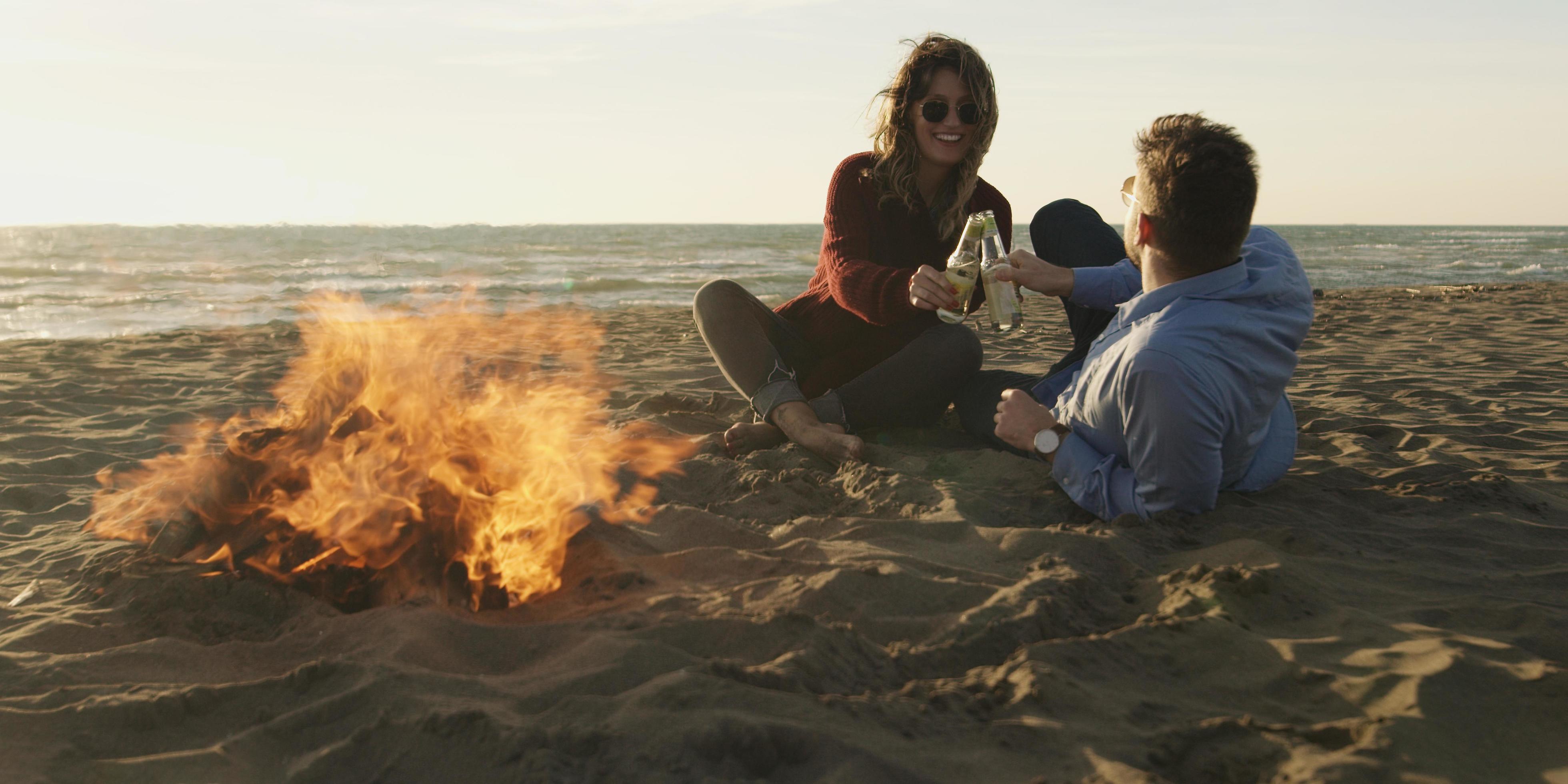 Loving Young Couple Sitting On The Beach beside Campfire drinking beer Stock Free