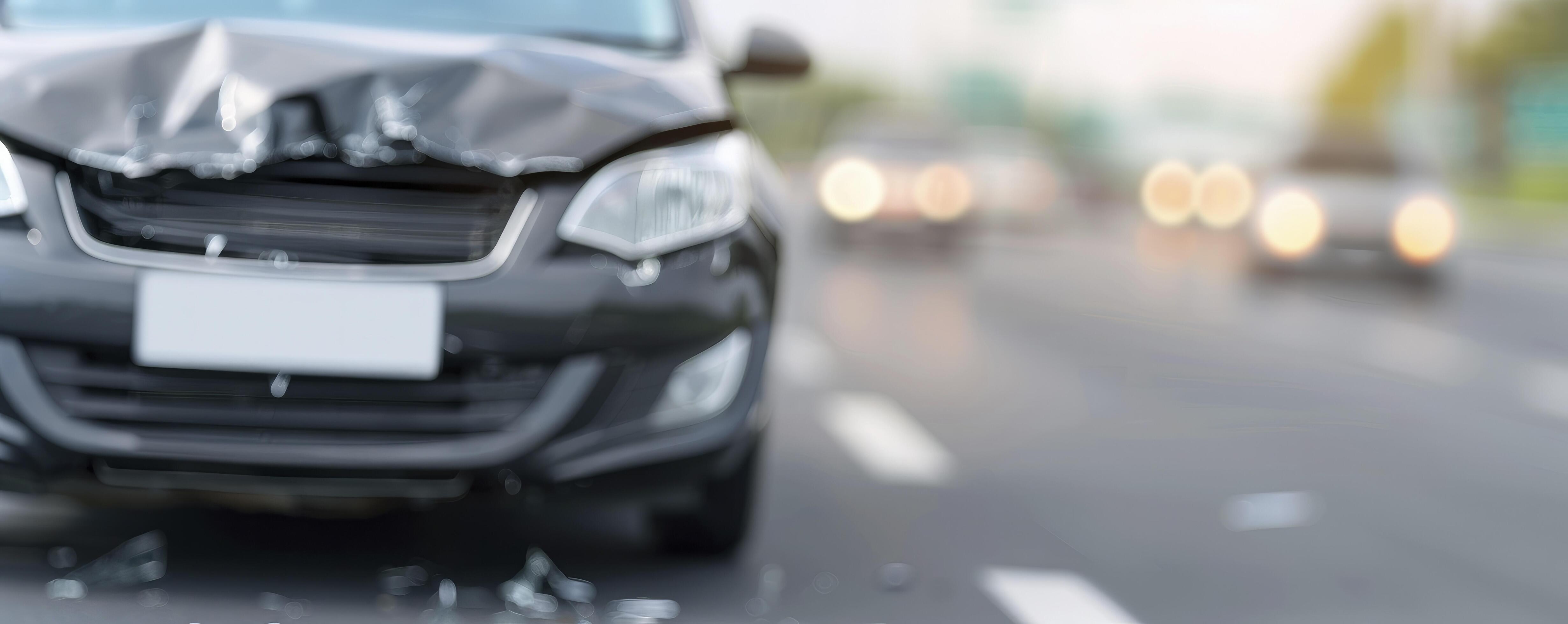 An accident damaged black car on the side of a highway, with cracks in its hood and headlights. The background shows other cars moving past. Stock Free