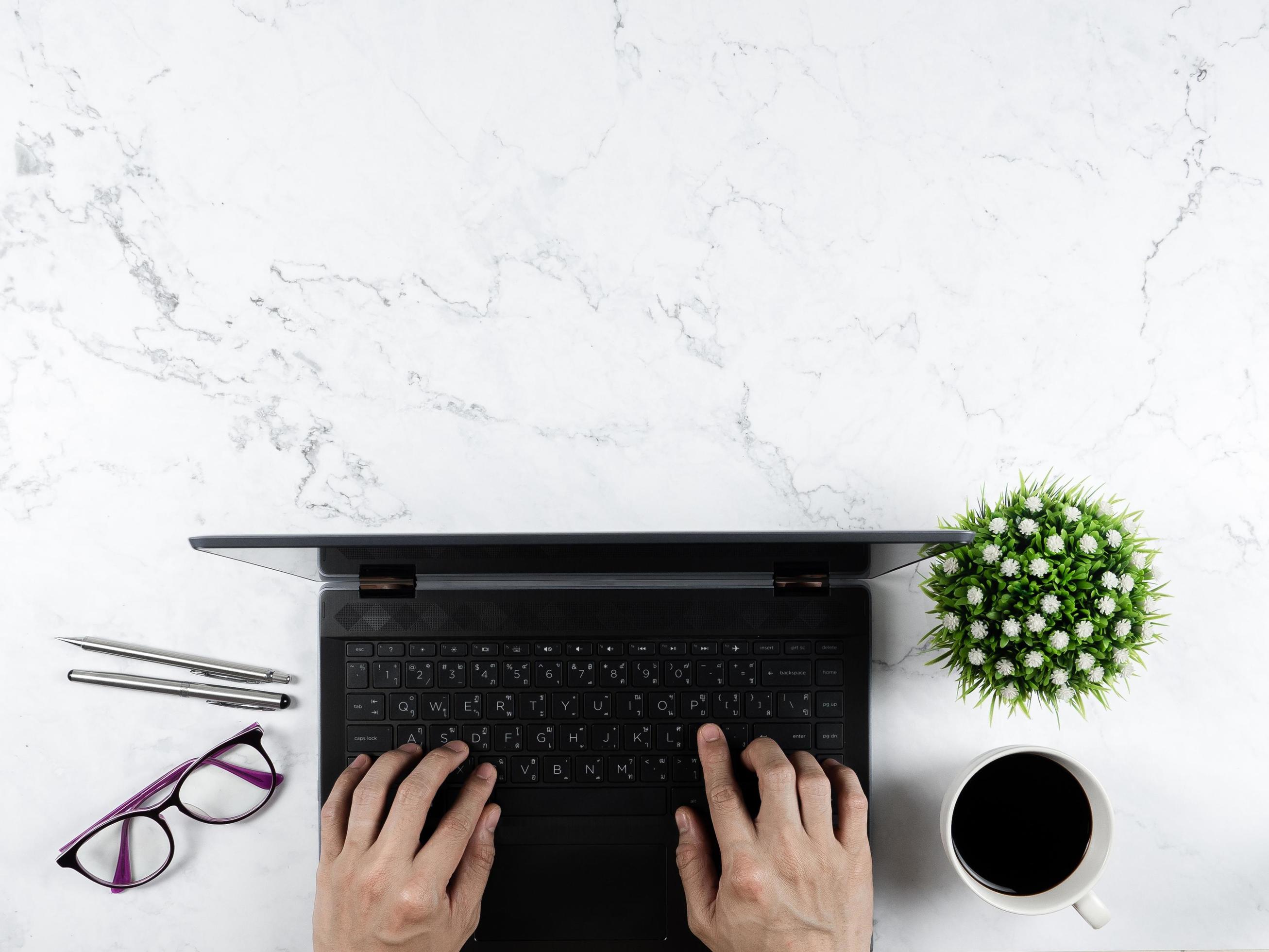 Hand on keypad laptop with glasses silver pen coffee cup flower vase on marble table workplace concept copy space top view Stock Free